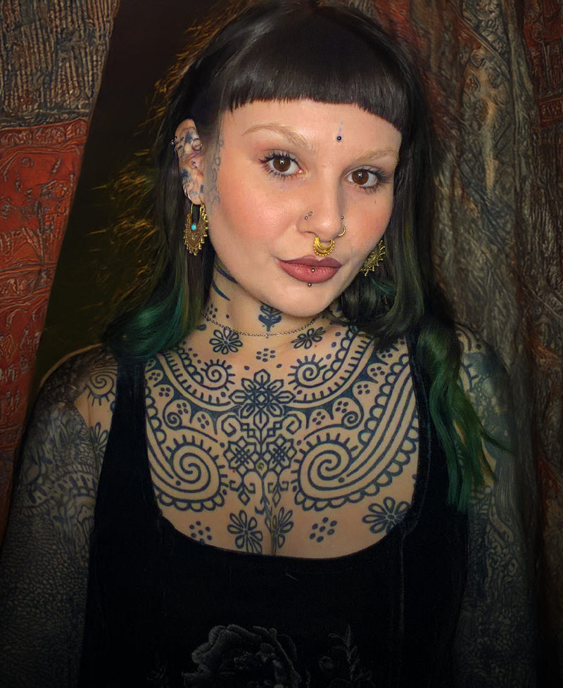 bohemian woman with tattoos, tribal earrings and piercings in black dress standing in front of Indian drapes