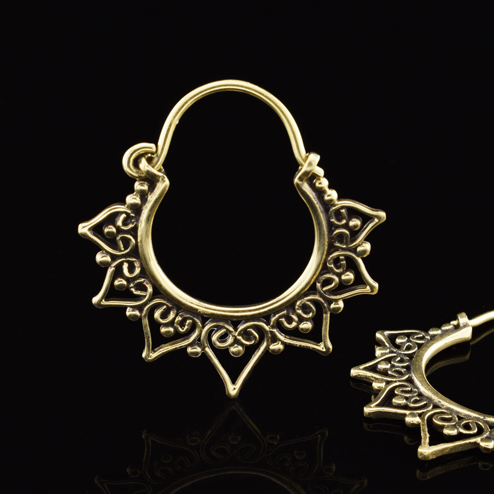 Golden Brass tribal hoop earrings with ornate heart filigree and pointed decorative ends, shown on a black reflective background with one earring fully visible and part of a second earring visible at the lower right.
