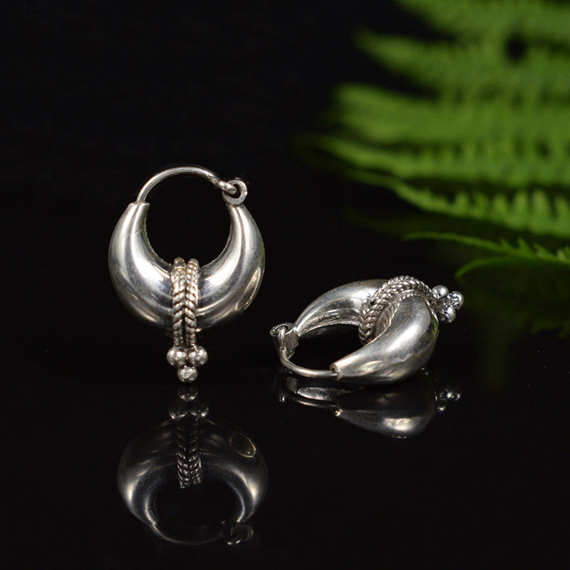 A pair of sterling silver crescent-shaped inidan hoop earrings with textured braided details and small bead drops, displayed on a reflective black surface with a green fern leaf in the background.