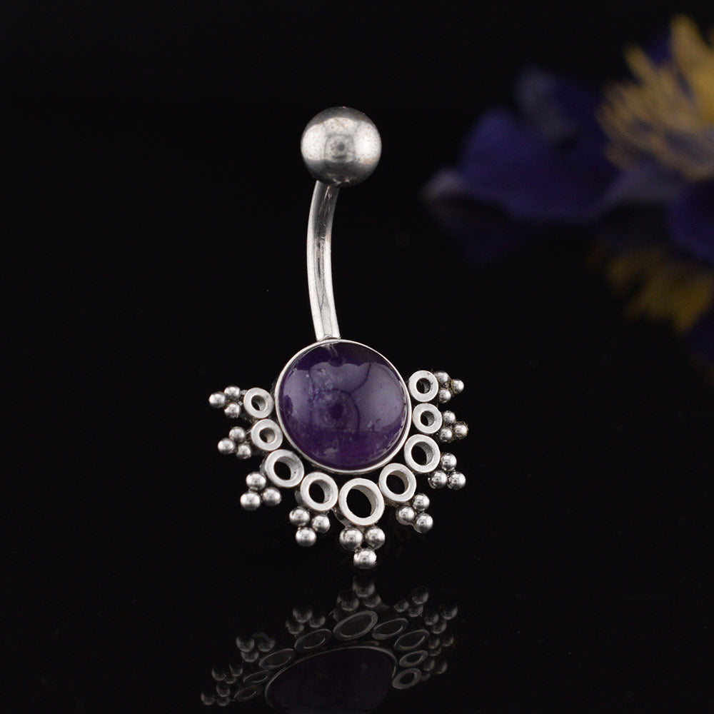 A tribal style navel bar / belly button ring featuring a round purple amethyst gemstone in the center, surrounded by a decorative cluster of small beads and openwork circles, set against a dark reflective background with blurred purple flowers.