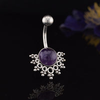 A tribal style navel bar / belly button ring featuring a round purple amethyst gemstone in the center, surrounded by a decorative cluster of small beads and openwork circles, set against a dark reflective background with blurred purple flowers.
