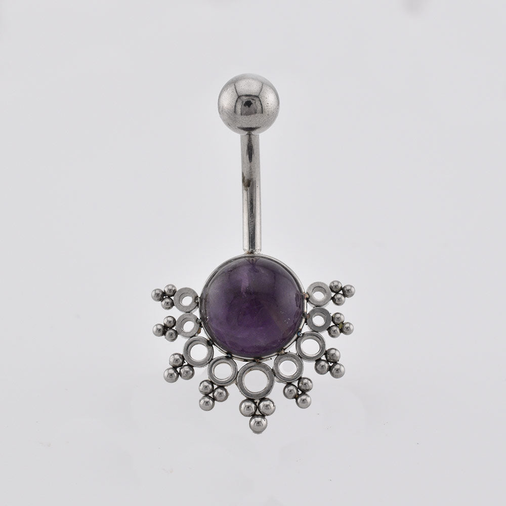 A navel piercing bar featuring a round purple amethyst in the center, surrounded by an intricate openwork silver filigree design, on a plain light gray background.