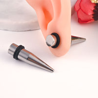A pair of steel tapered ear plug spikes with black rubber rings, shown with one lying on a white surface and the other inserted into a peach-colored silicone ear model.