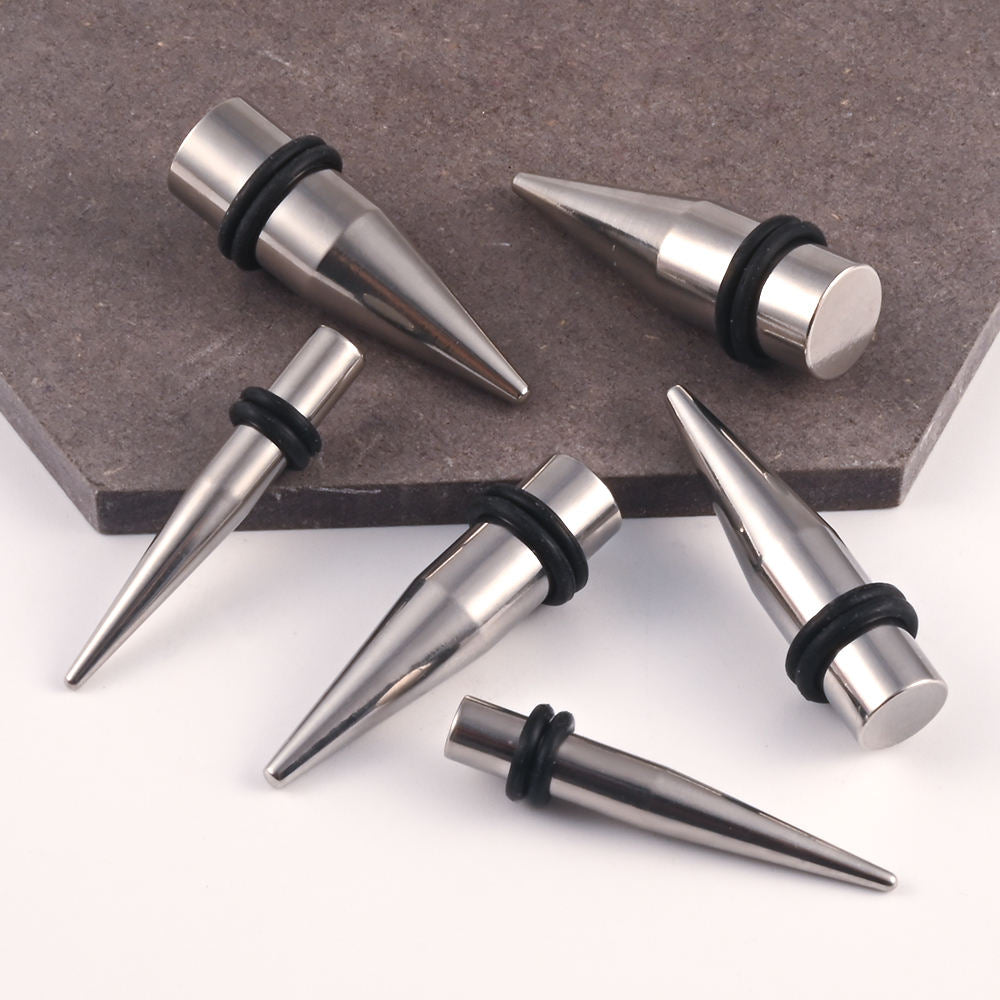 Five steel ear stretching spike plugs  with black rubber O-rings, arranged on a white surface with a dark gray stone tile in the background.
