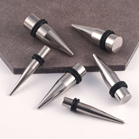 Five steel ear stretching spike plugs  with black rubber O-rings, arranged on a white surface with a dark gray stone tile in the background.