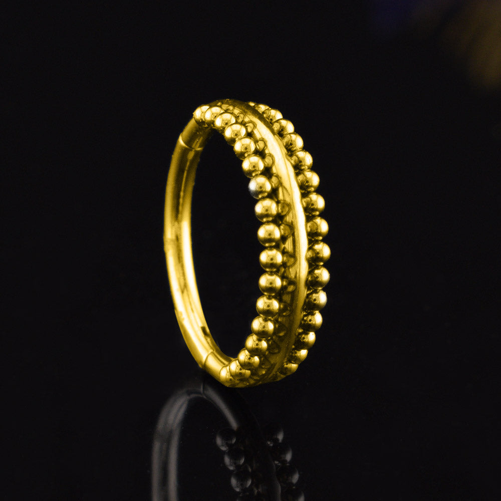 A gold steel hinged segment ring with a double row of small, polished bead-like details, shown on a reflective black background.