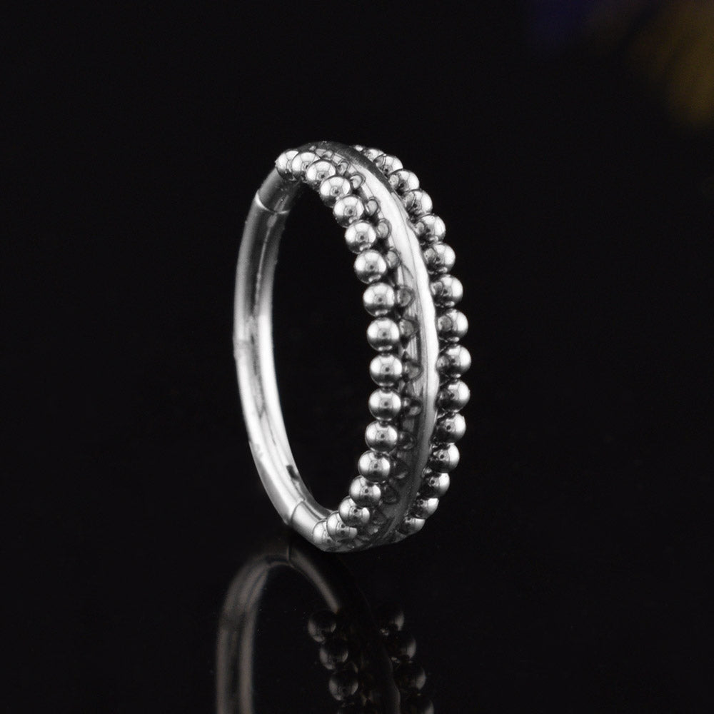 A surgical steel triple-band hinged segment clicker ring featuring one smooth band and two beaded bands, displayed on a reflective black surface.