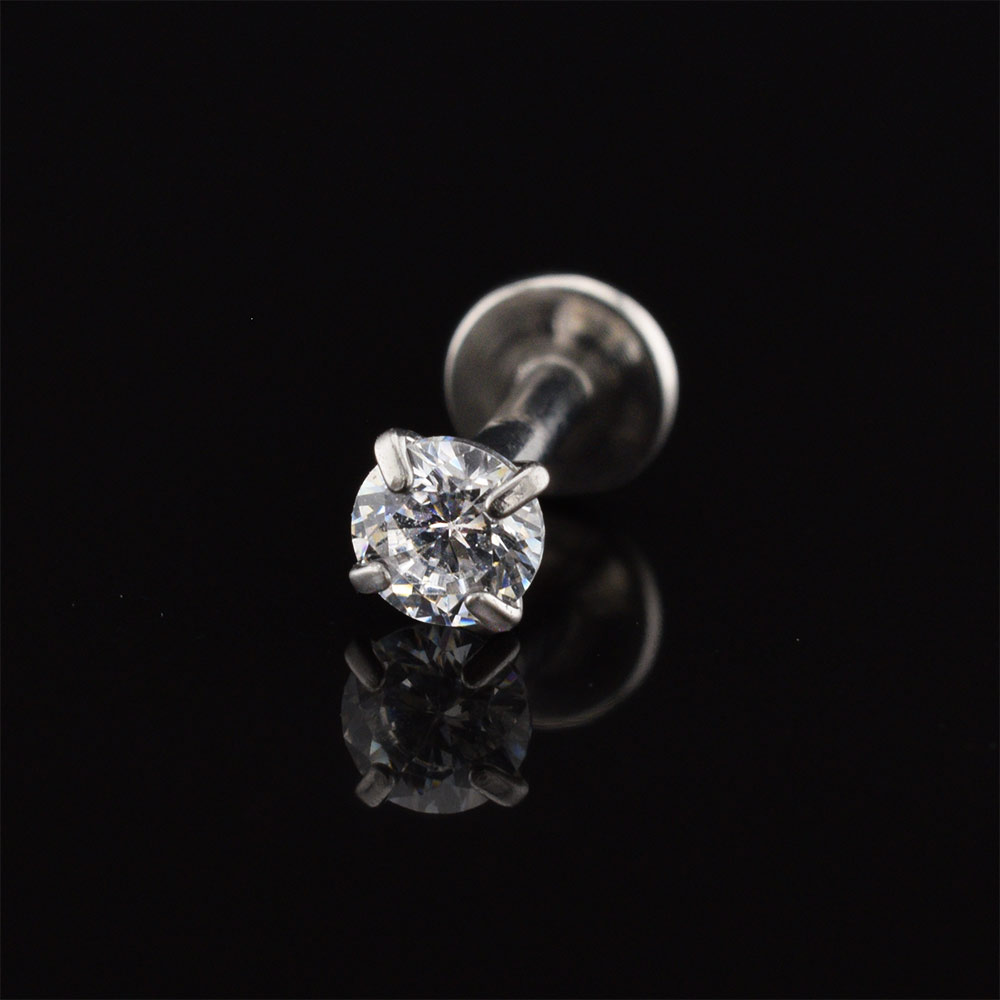 A single solitaire labret stud piercing jewelry piece featuring a round brilliant-cut clear gemstone in a four-prong setting on a silver-toned post and flat-back postback, displayed on a reflective black surface.