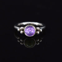 A surgical steel hinged segment clicker ring featuring a round purple amethyst gemstone in the center, flanked by small silver bead details on the shoulders, set against a solid black background with a soft reflection below.