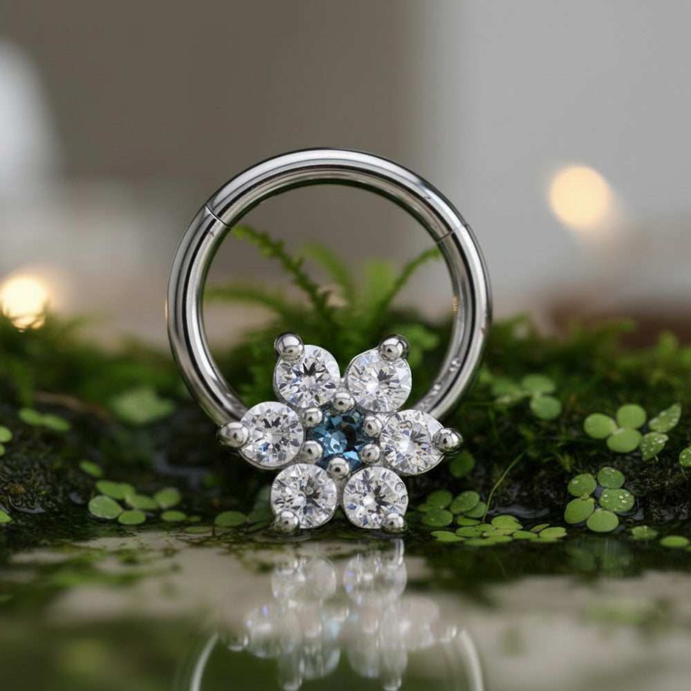 A hinged segment clicker earring with flower design in CZ crystals in a flower-shaped cluster of clear gemstones and a small blue center stone, photographed resting on green moss with a soft blurred background.