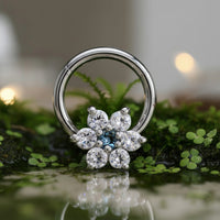 A hinged segment clicker earring with flower design in CZ crystals in a flower-shaped cluster of clear gemstones and a small blue center stone, photographed resting on green moss with a soft blurred background.