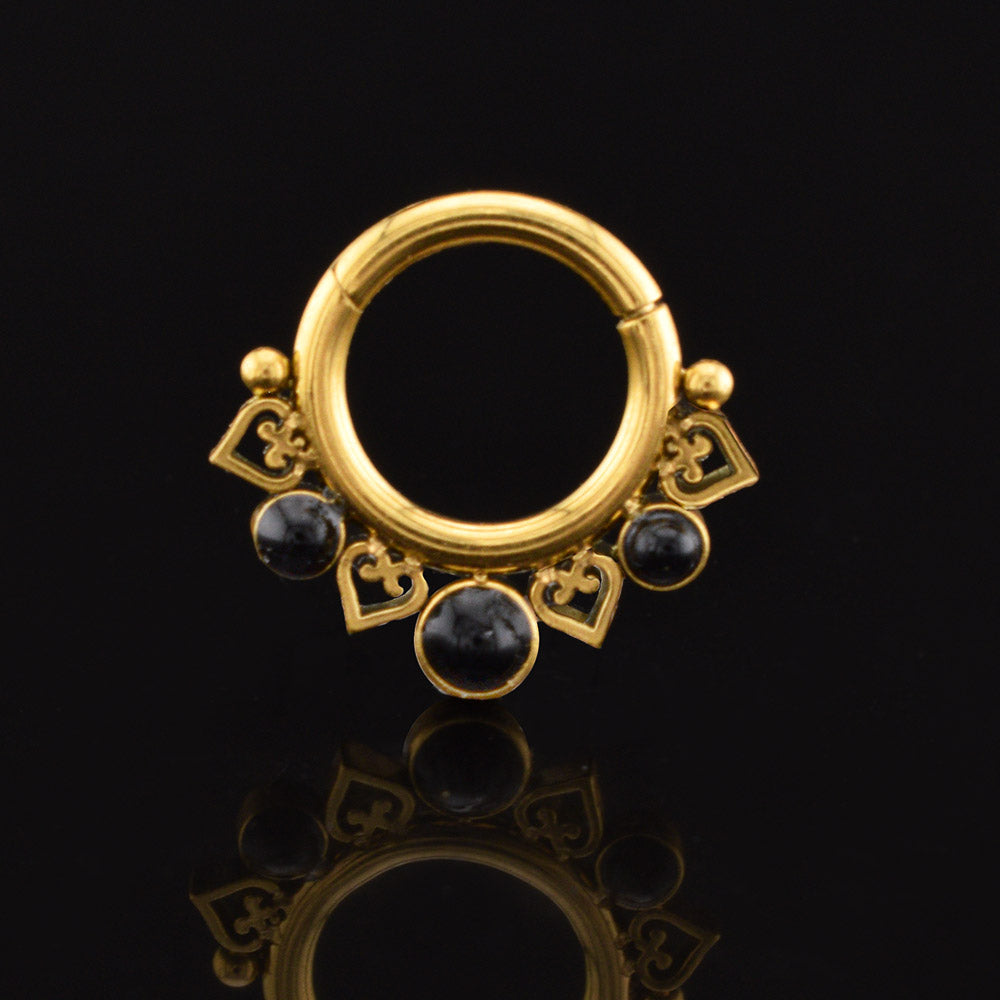 A gold pvd clicker ring piercing with decorative cutout heart and circle details, accented by three small round black onyx stones, shown on a reflective black background.