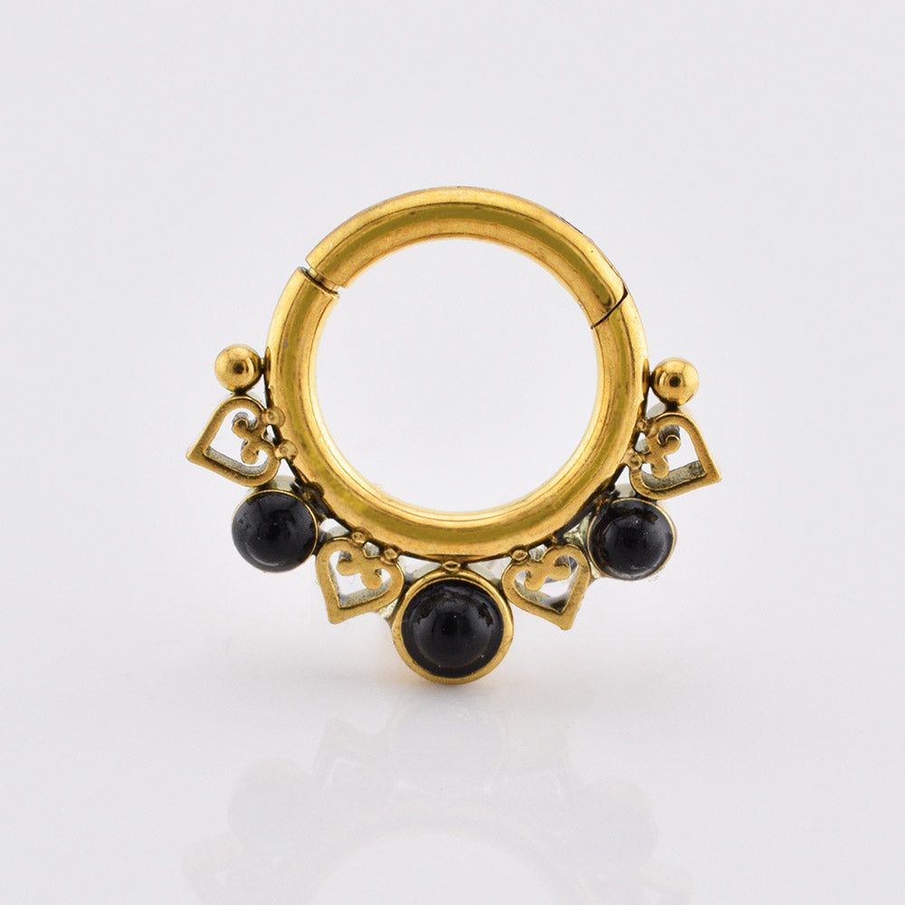 A gold pvd hinged segment ring piercing with decorative cutout heart and circle details, accented by three small round black onyx stones, shown on a white background.