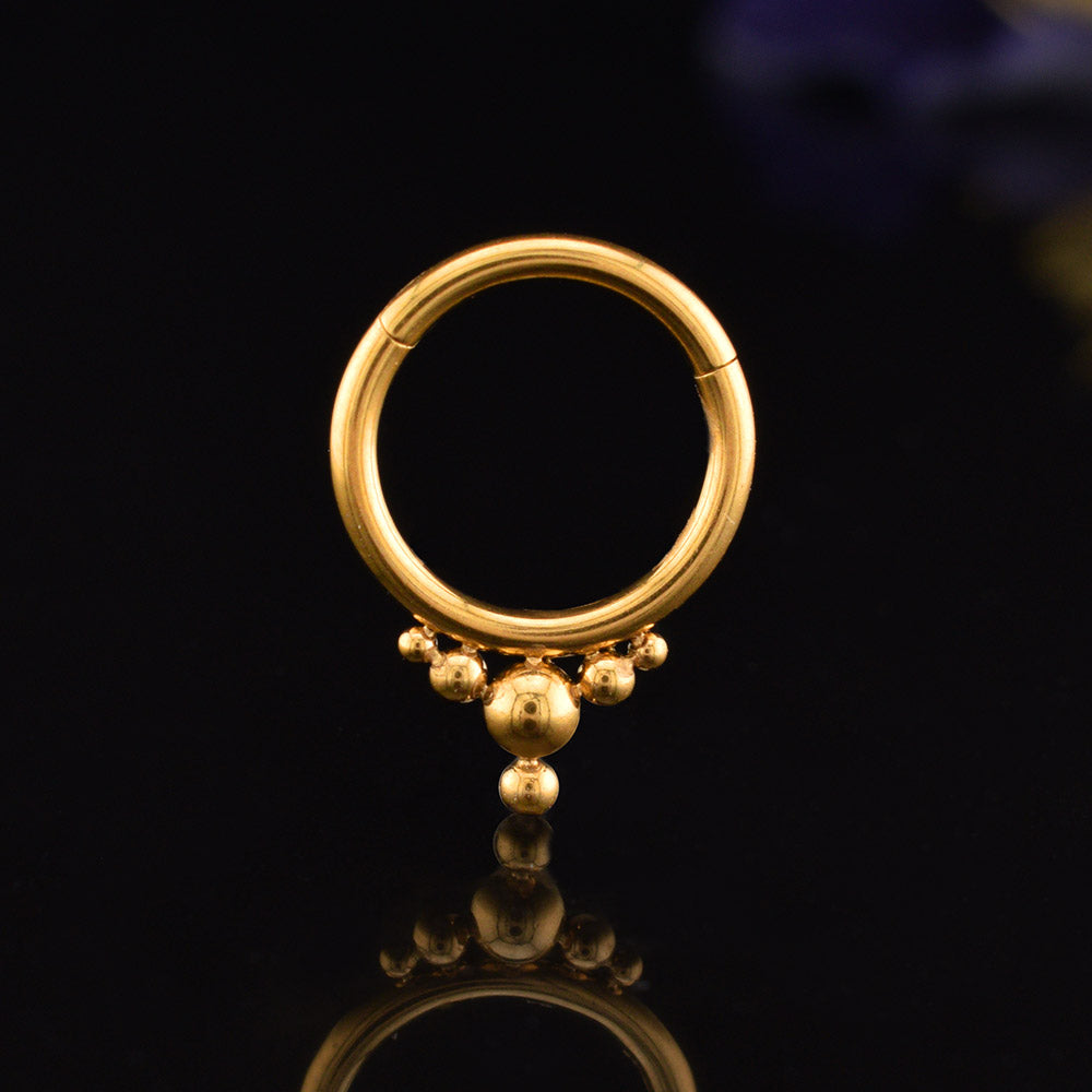 A gold  steel hoop hinged segment piercing ring with a fixed tribal beaded fringe, displayed on a reflective black surface.